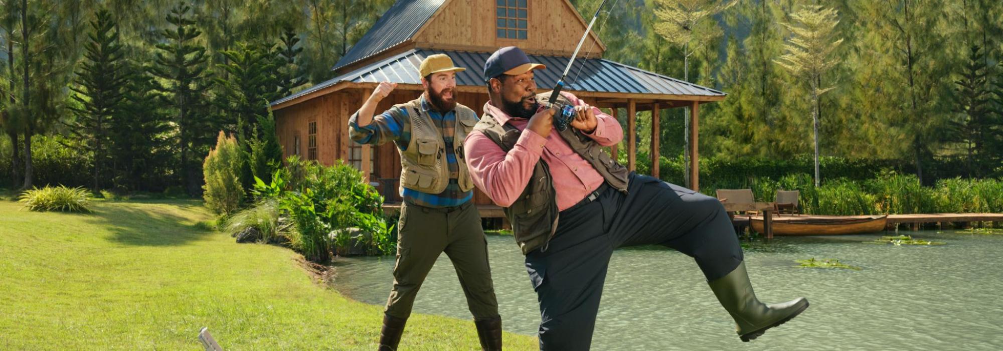 Two men by a cabin next to a pond, one holds a fishing rod