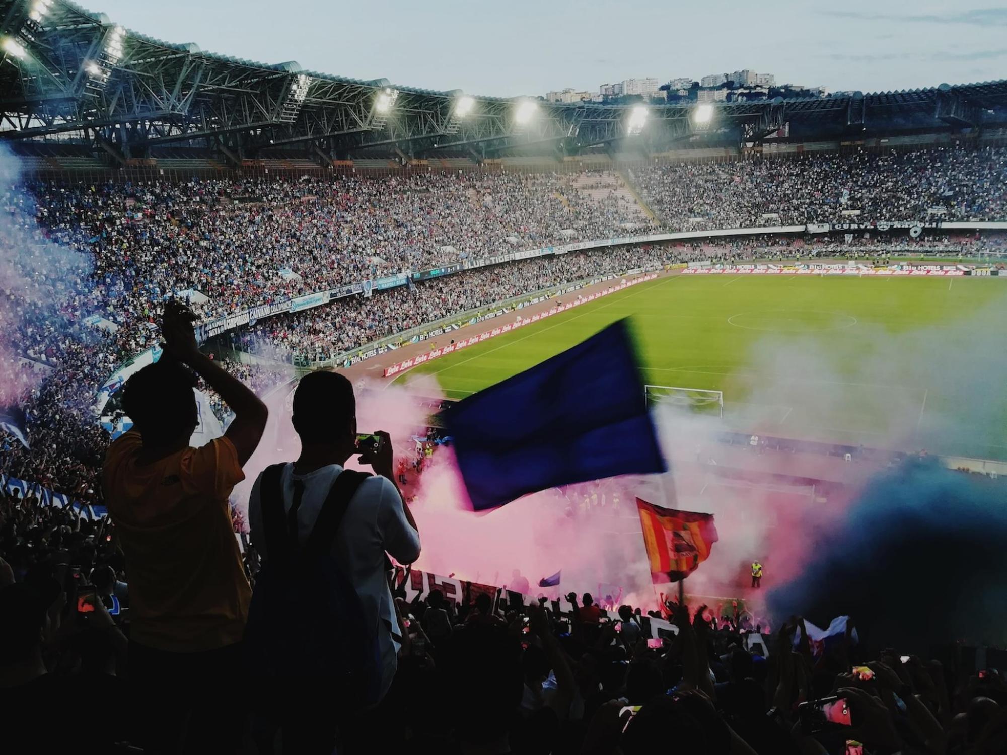 Fans in a stadium waving flags and lighting flares