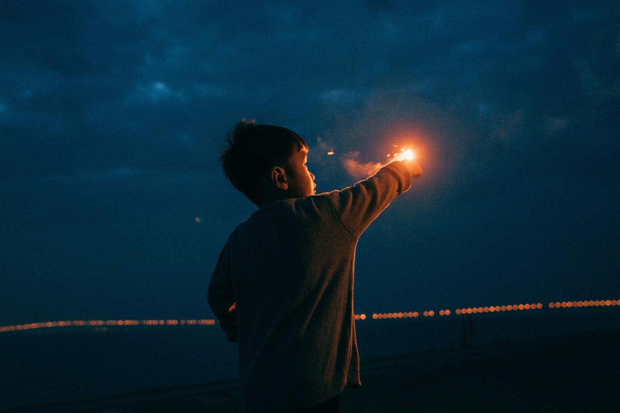 A boy stands in darkness, his face illuminated by a sparkler