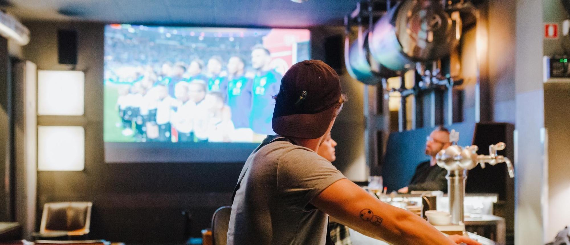 A man stands at a bar in a sports bar in NYC watching the TV