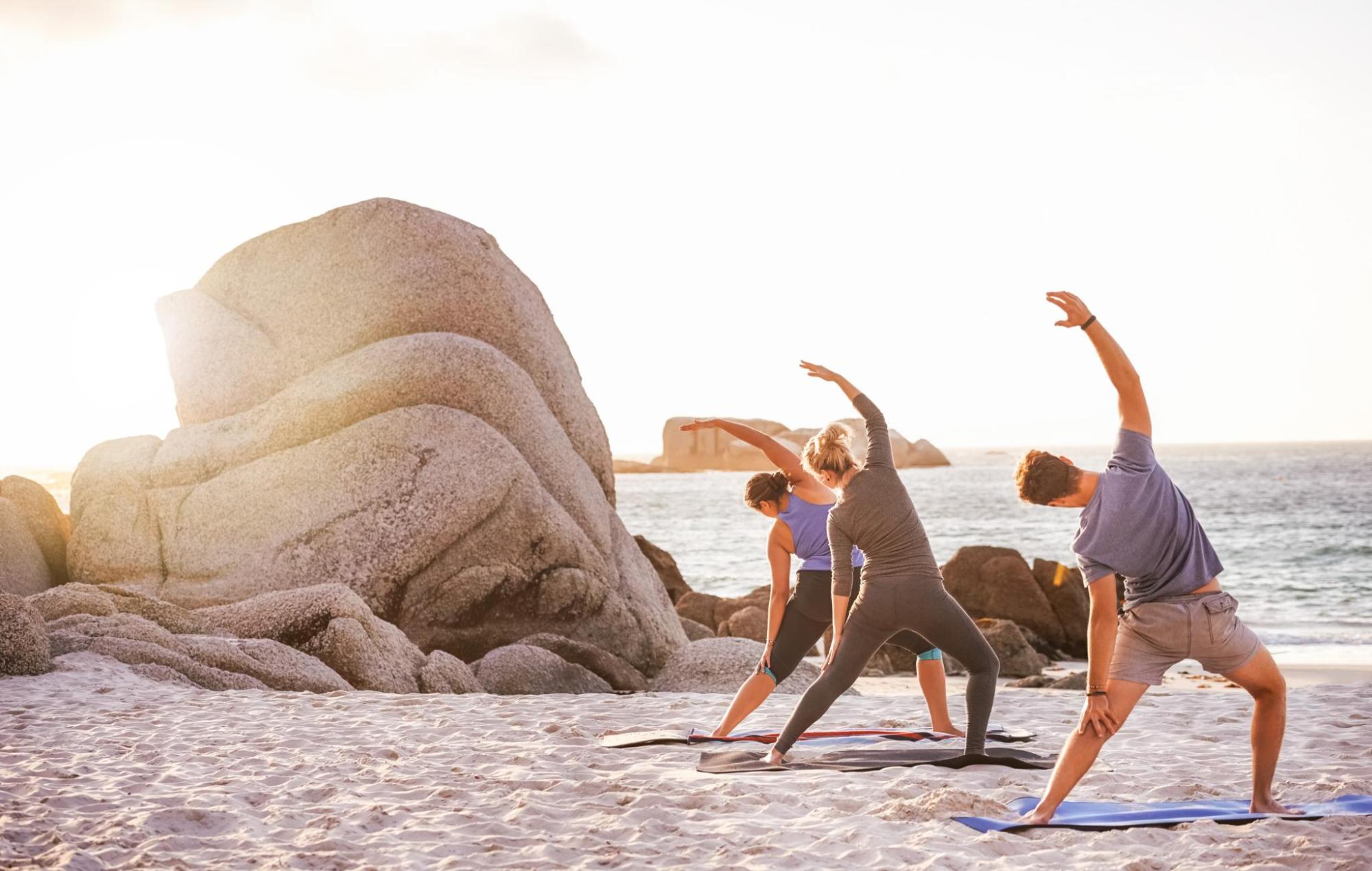 Three individuals practise Pilates poses on a sandy beach.