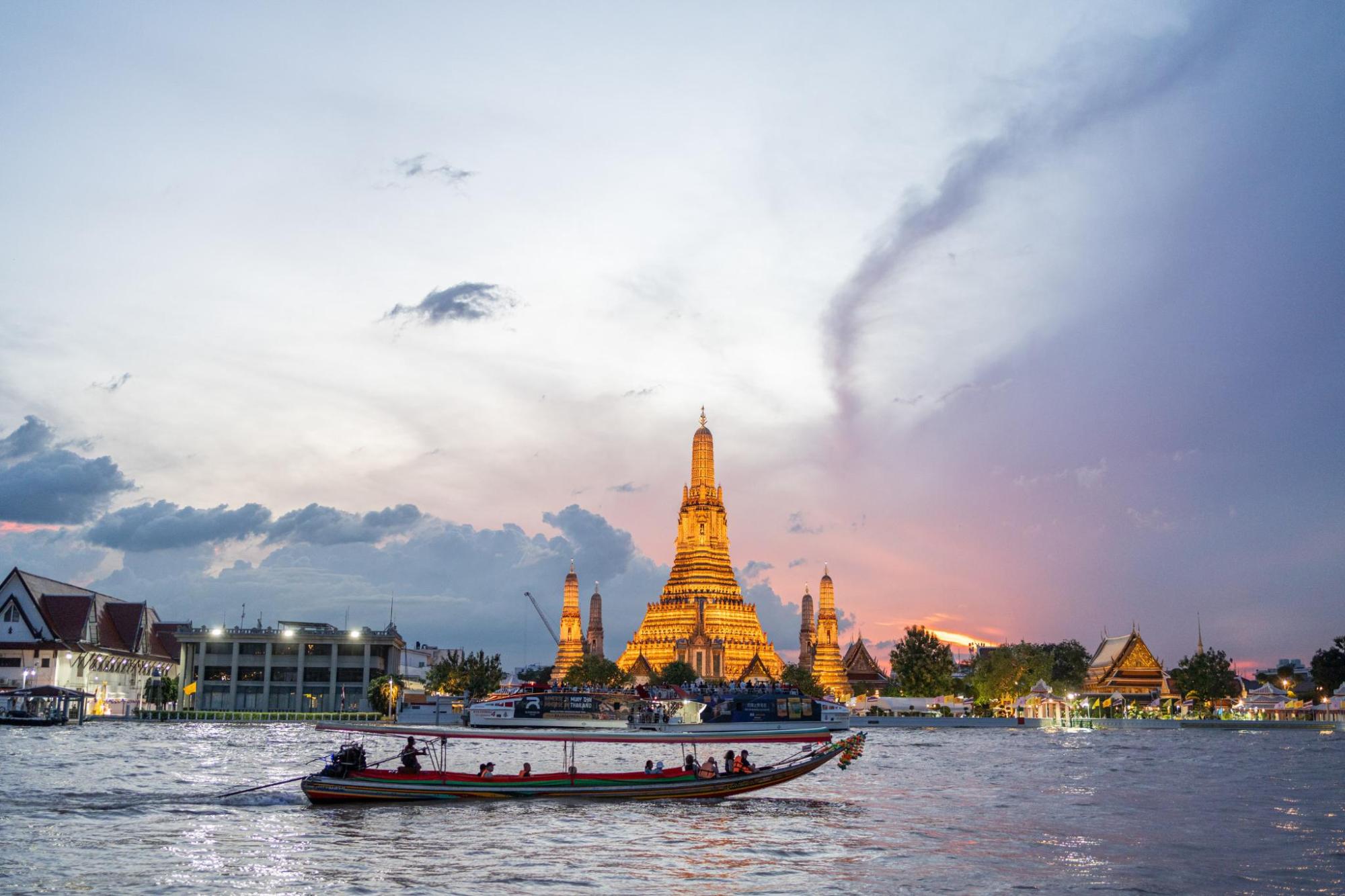 Wat Arun temple lit at sunset across river in Bangkok