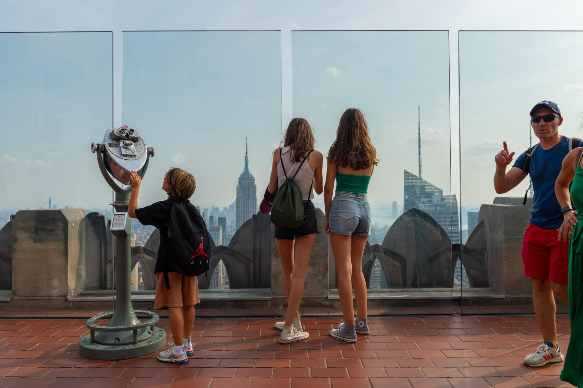 Two teens view NYC's cityscape, a boy looks at a telescope