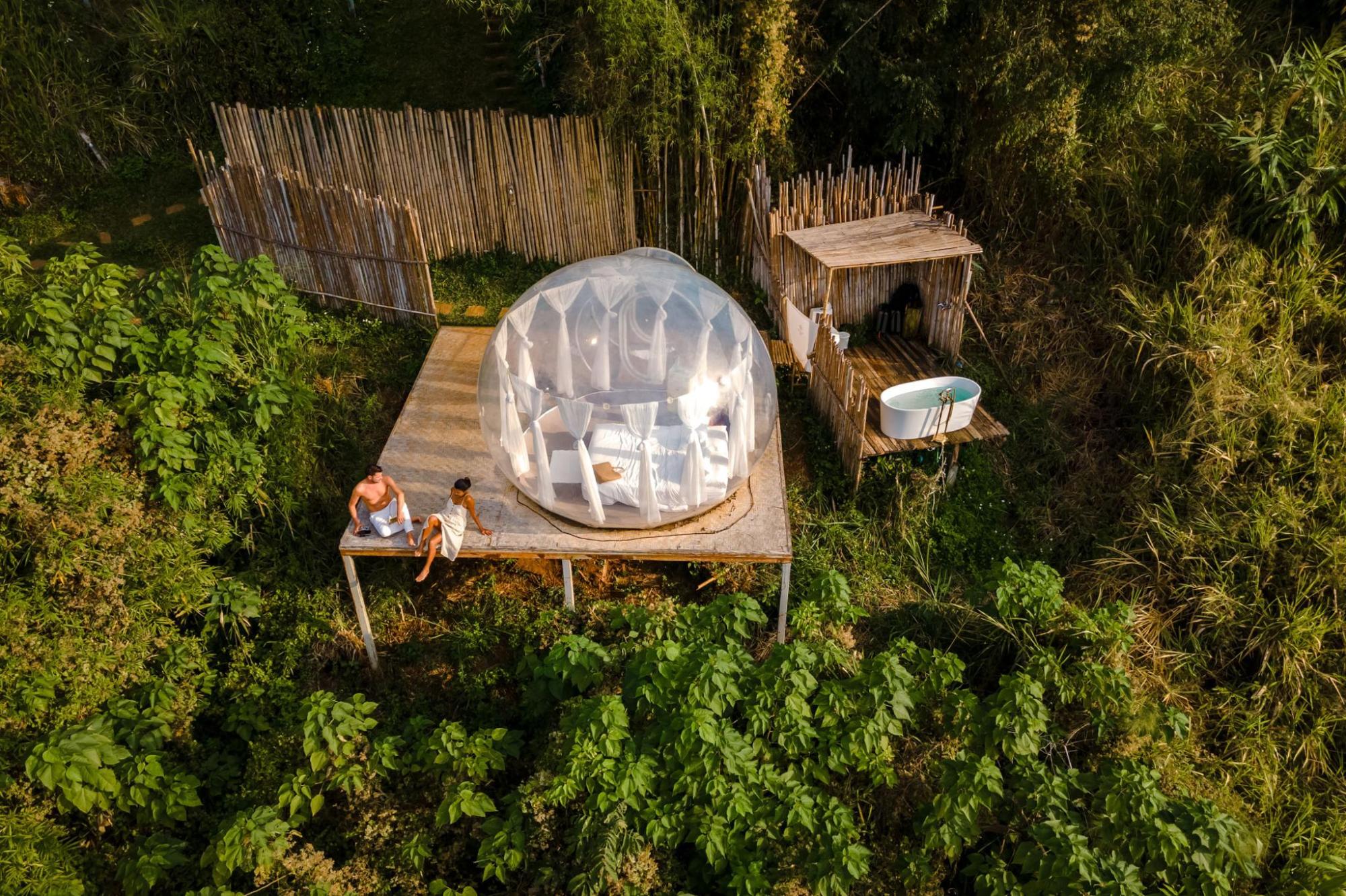 A couple sits next to a transparent dome in the middle of a forest, offering a unique view of the surrounding