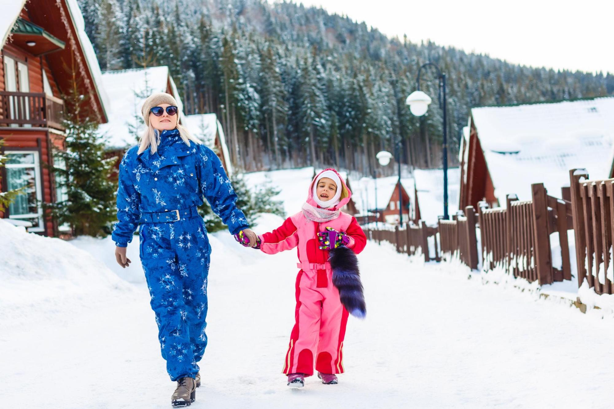 A woman holding the hand of a young girl whilst they walk in the snow in front of some wooden cabins