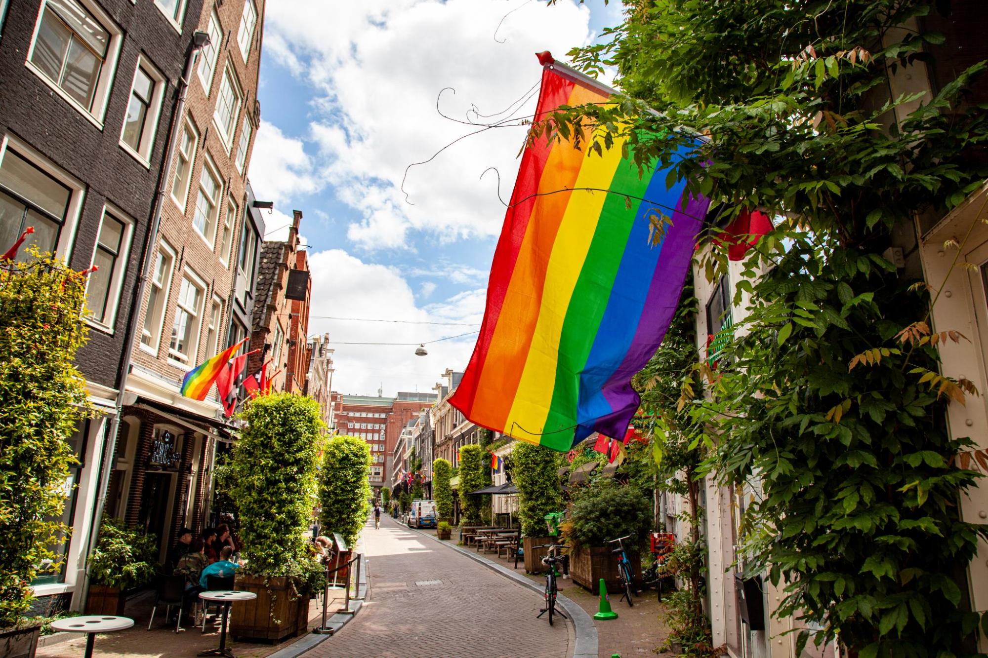 Rainbow flags hang above a narrow street lined with cafés.