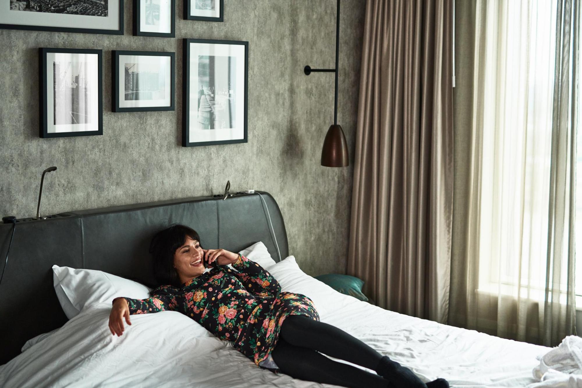 A woman in a floral dress talks on a phone while lying on a bed in a modern hotel room