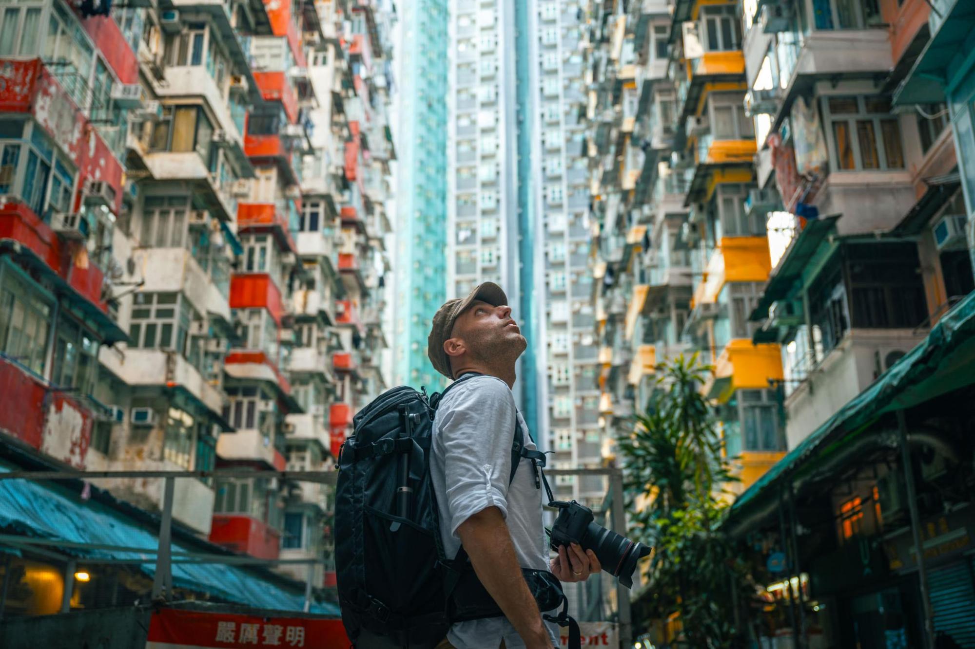 A person with a backpack looks up at tall buildings in a narrow alleyway