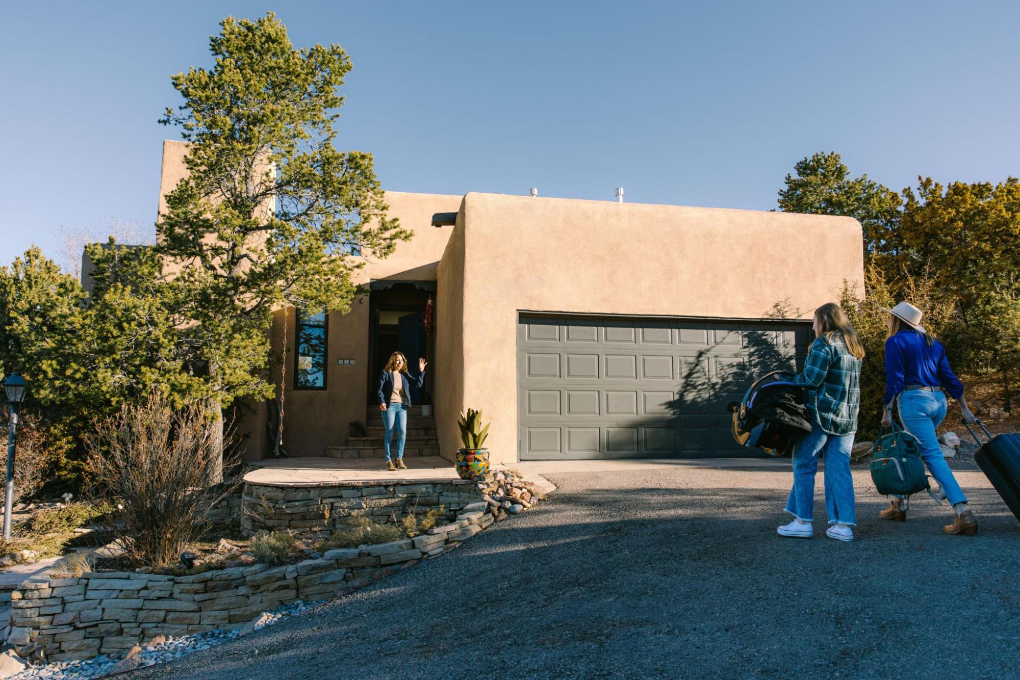 A homeowner stands outside a large brown house, warmly welcoming two guests.