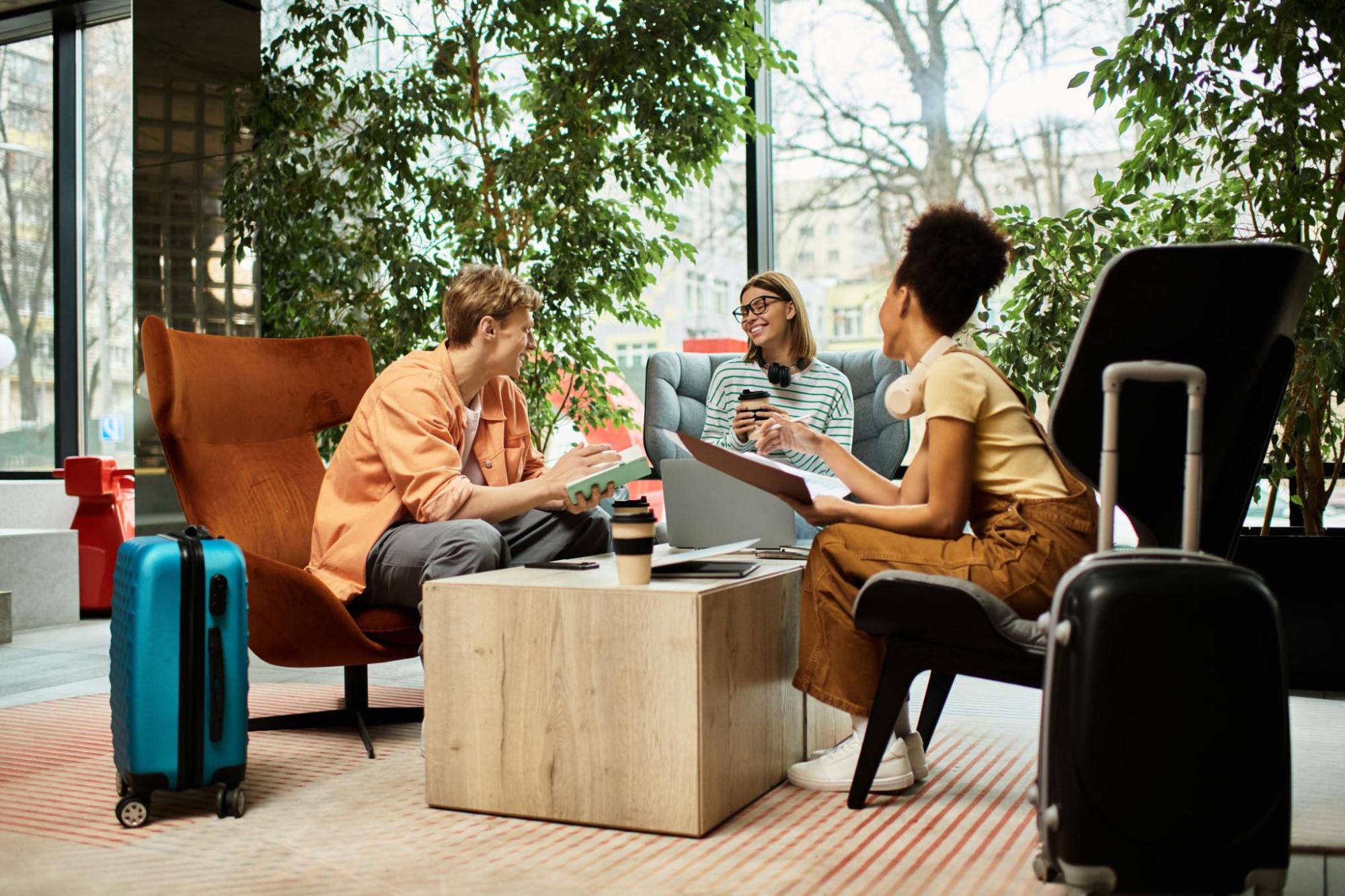 Three travellers chat around a low table in a bright lounge, with suitcases, coffee cups, a laptop and large plants by the windows