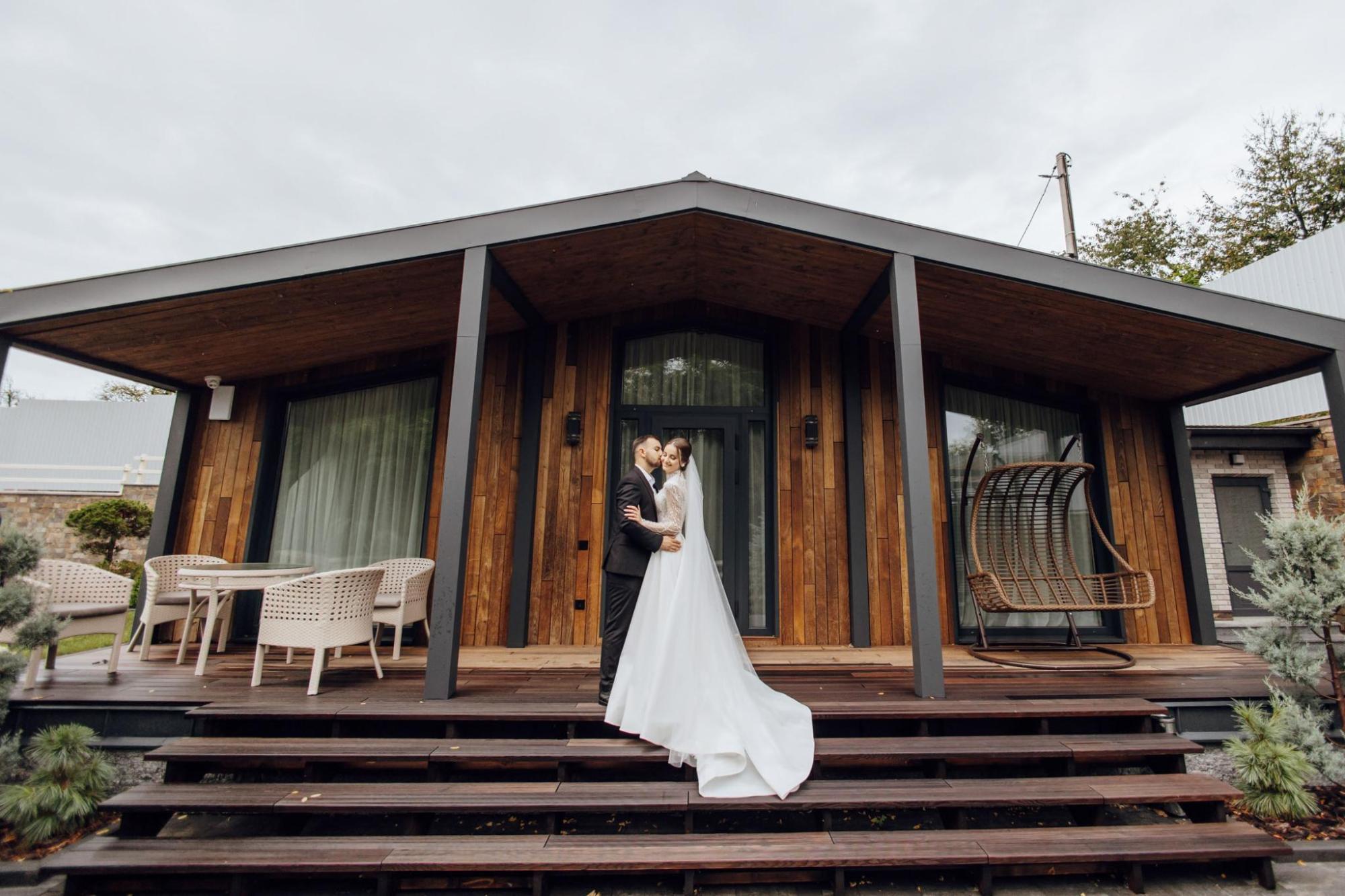 A bride and groom stand on the steps outside a modern wooden cabin with a covered terrace, outdoor seating and large windows