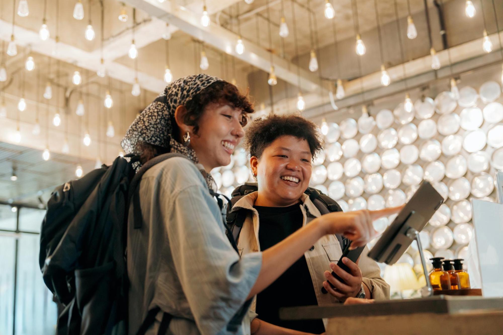 Two smiling travellers with backpacks check in at a reception desk, looking at a screen beneath hanging lights in a stylish lobby