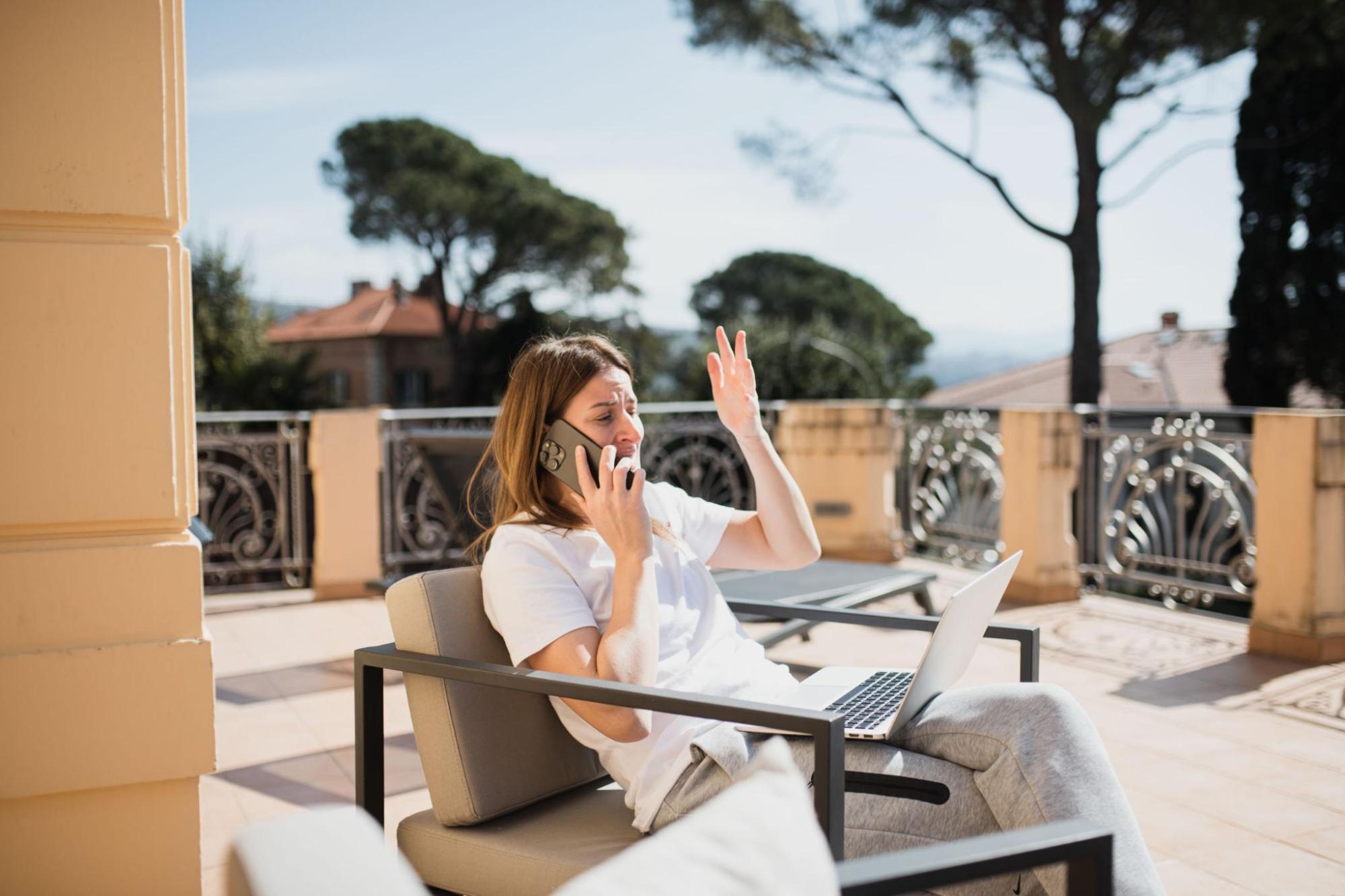 A traveller sits on a sunny terrace with a laptop, talking on a mobile phone and raising a hand, with trees in the background