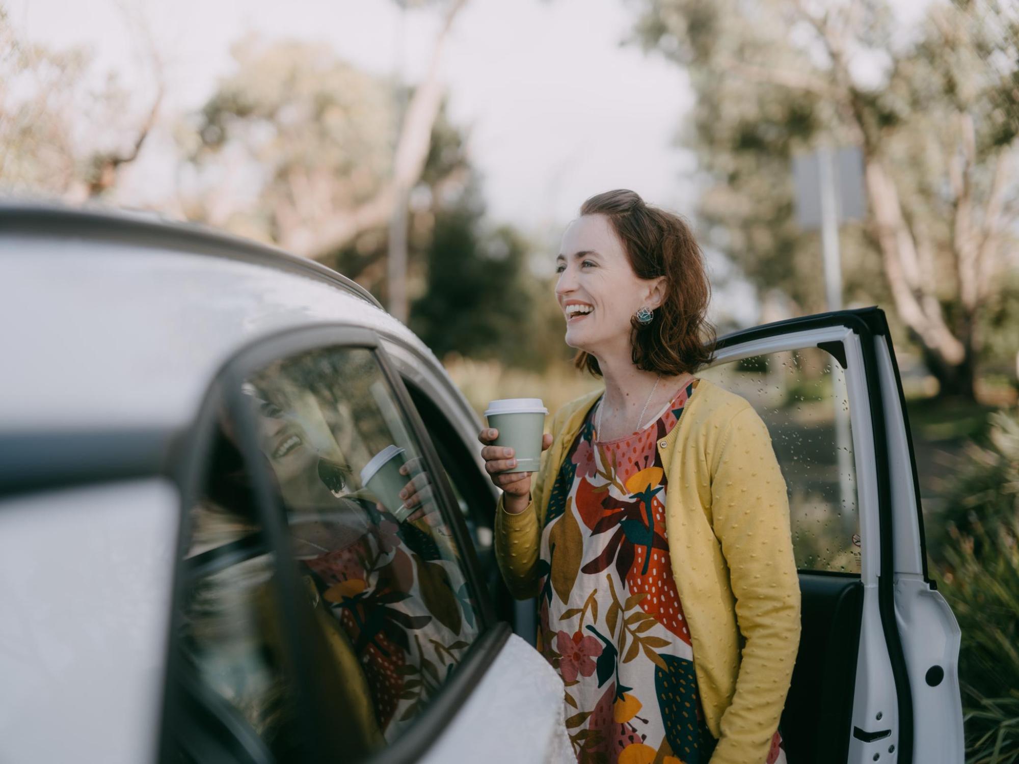 A person in yellow clothing stands beside a white car, holding a takeaway coffee