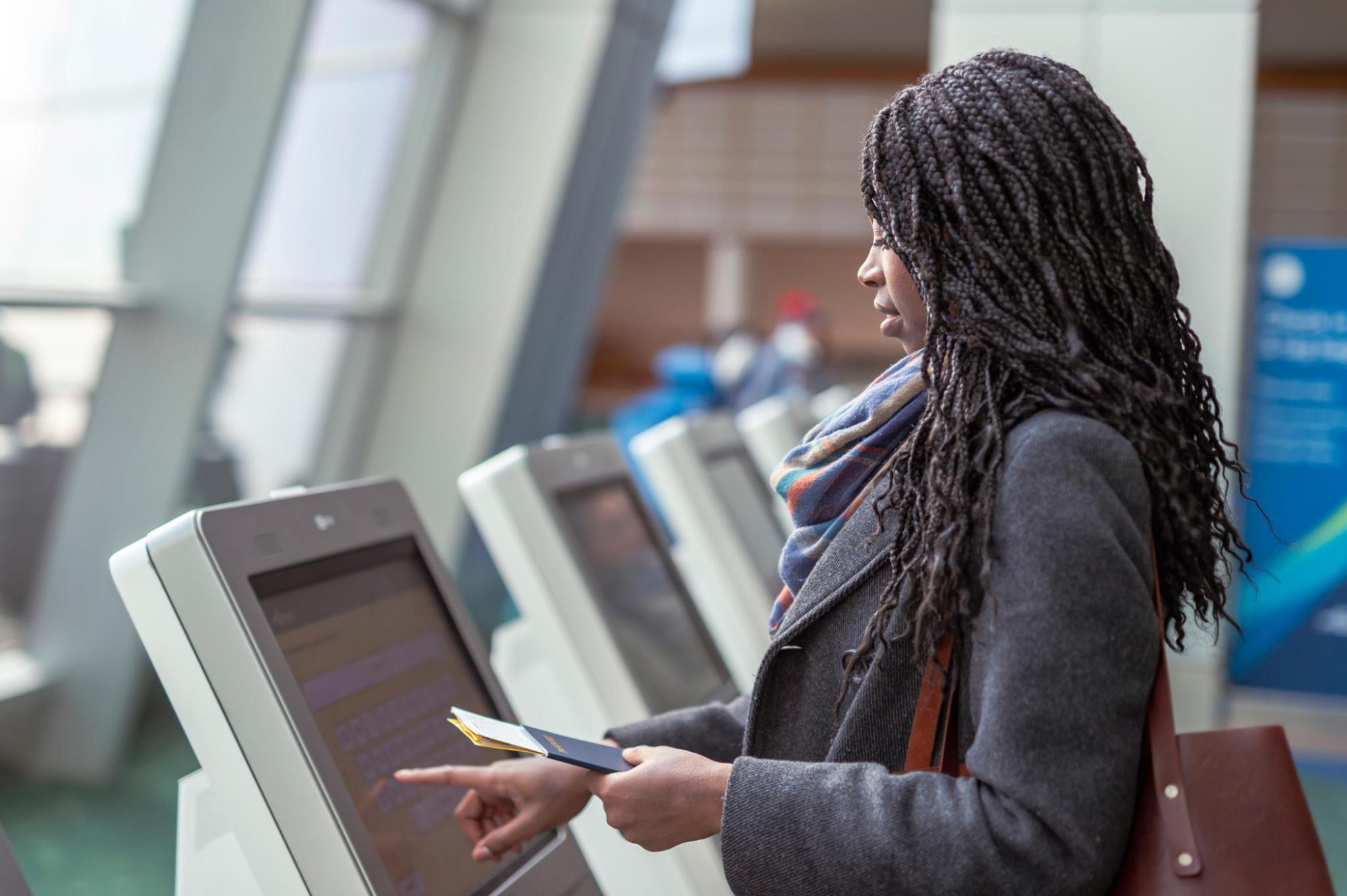 A person at an airport terminal uses a self-check-in machine, displaying their passport