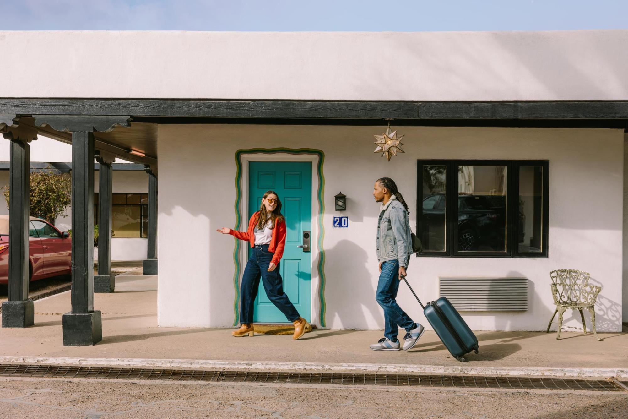 Two guests standing outside a motel room with a turquoise door, as one person rolls a suitcase along the walkway