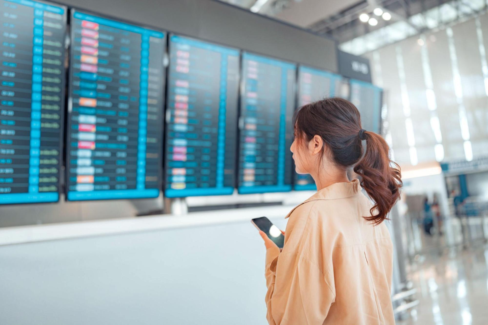 Woman holding a phone looks at flight information boards inside an airport terminal