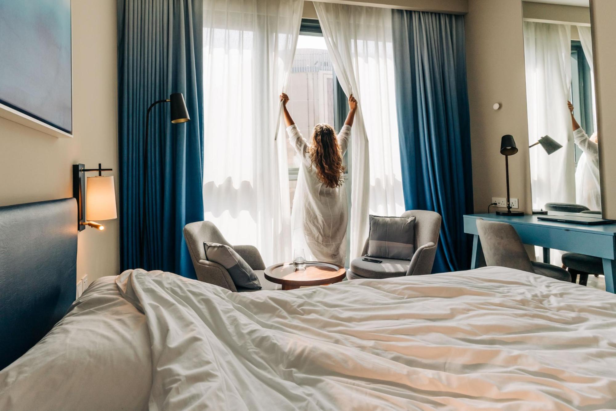 A person opening a curtain in a guest room in the morning light
