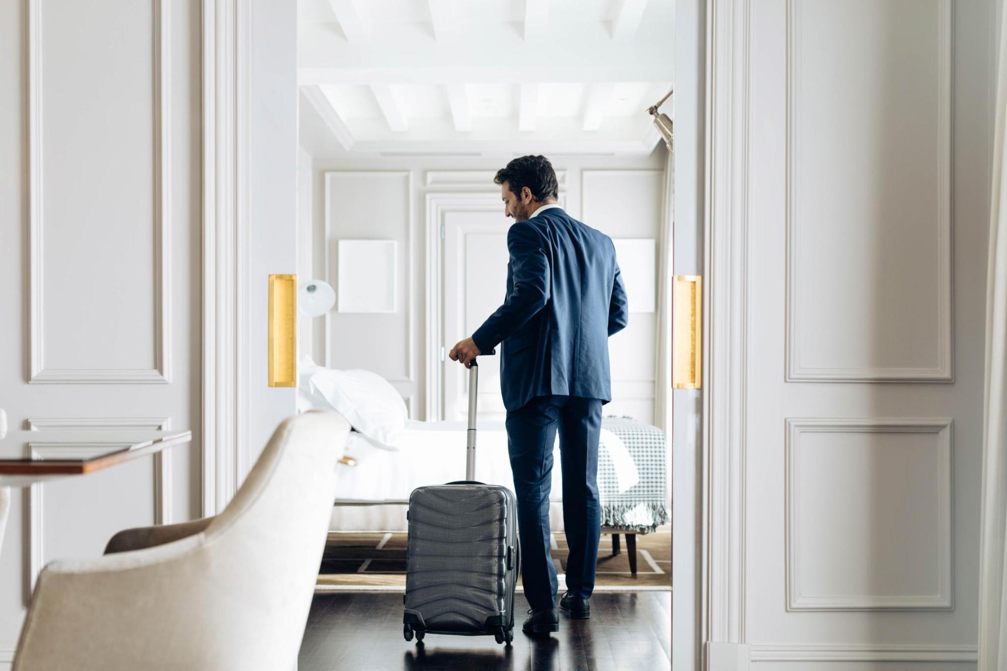 A business traveller in a suit walks through a guest room carrying a suitcase