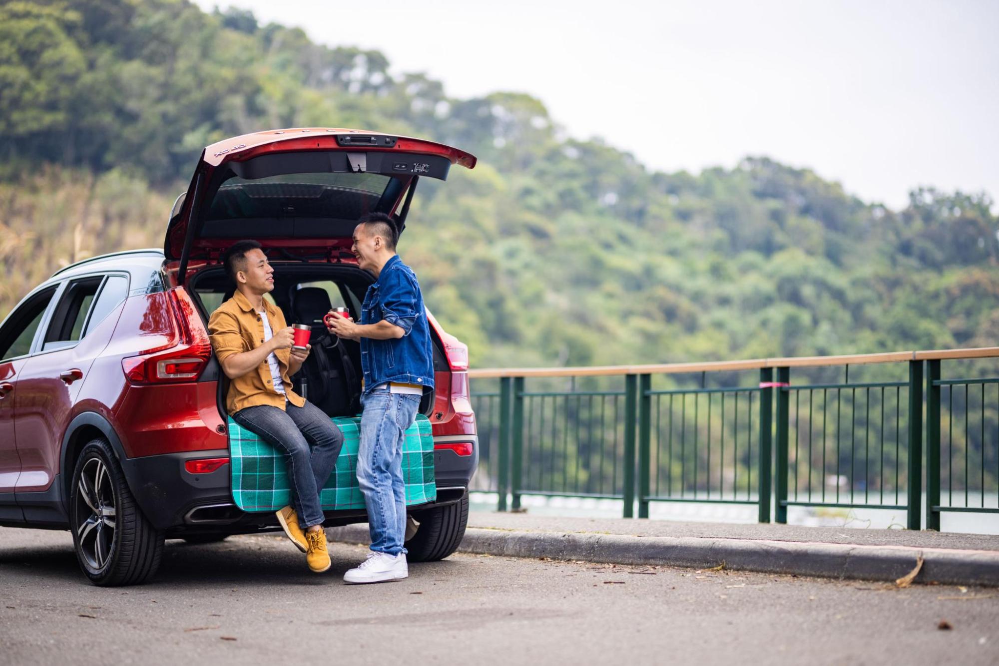 Two travellers chat at the open boot of a red SUV, holding cups beside a roadside lookout with forested hills in the background