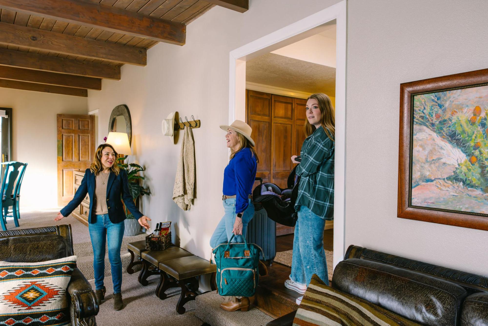 Host welcomes two guests with luggage into a cosy holiday home living room with wooden beams and warm, rustic décor