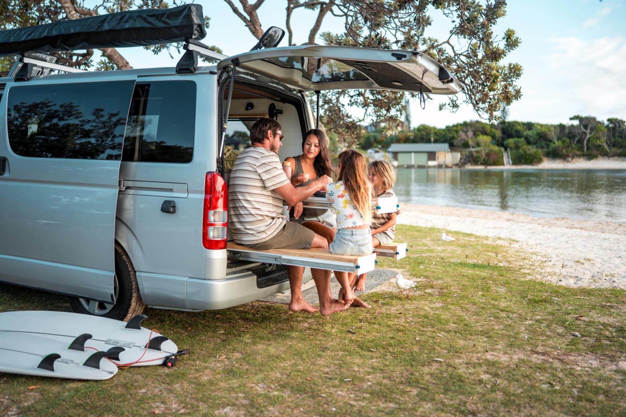 Two adults and two children sit on the back of a van beside a lake