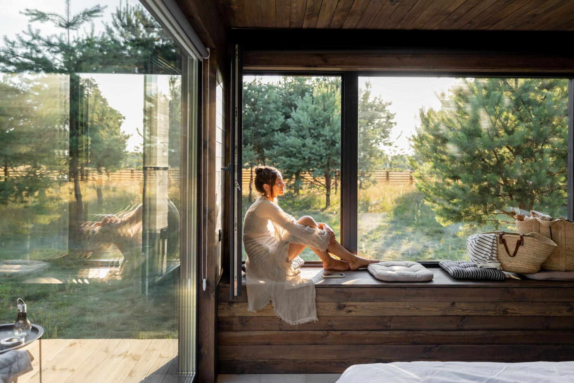Woman sitting on a window seat in a wooden cabin, looking out at pine trees in bright light