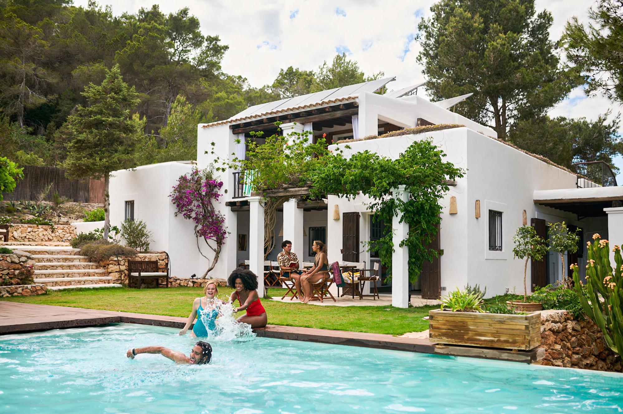 Group of three people swimming in a pool with a white villa in the background, with two persons sitting on the shaded terrace