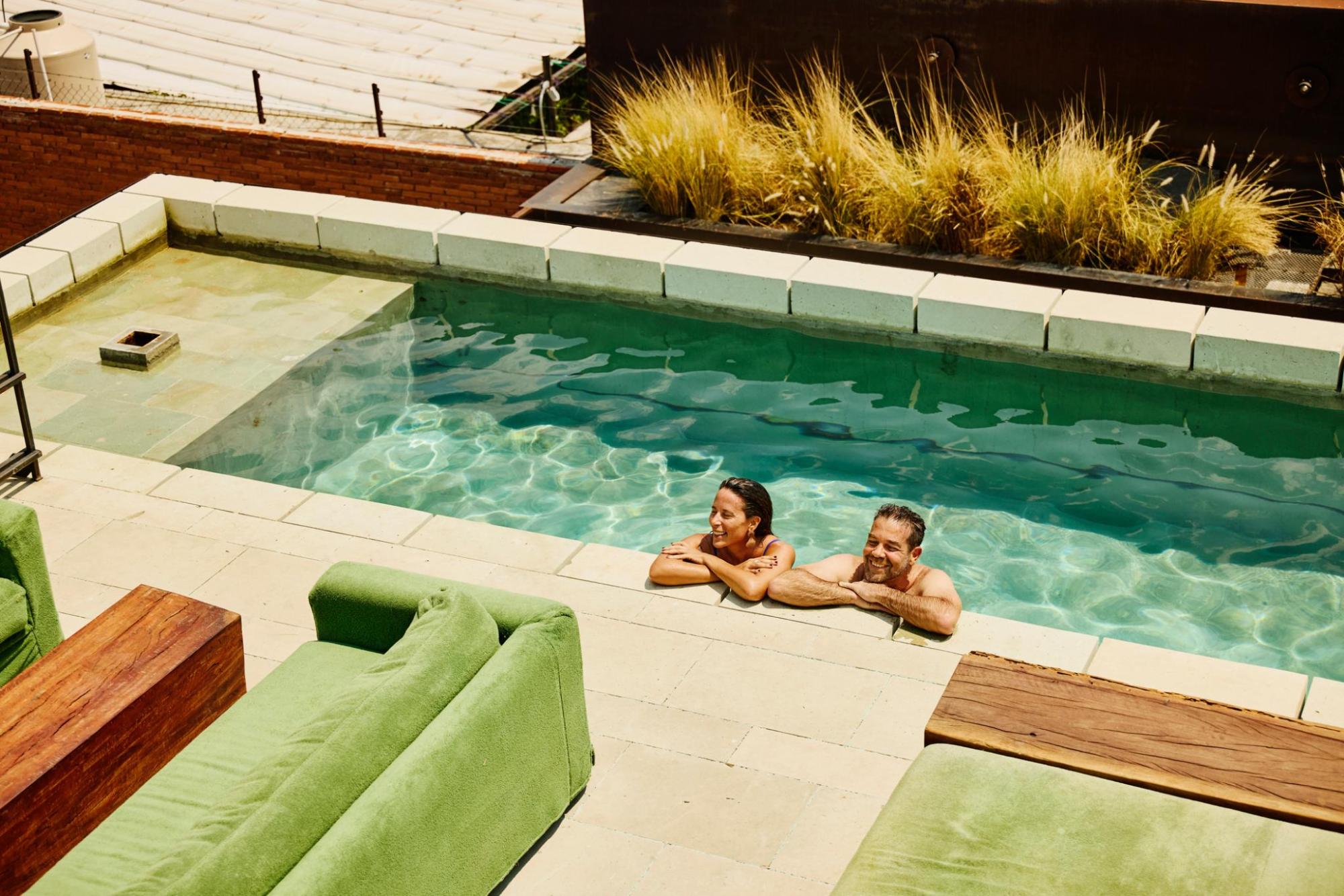 Two people smile while relaxing in a swimming pool next to green outdoor sofas