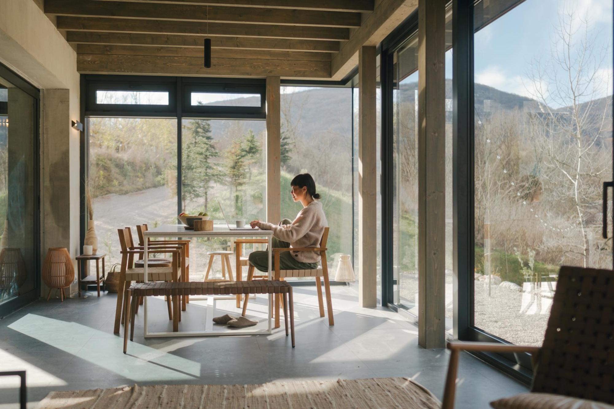 Woman working on a laptop at a dining table in a bright cabin with floor-to-ceiling windows and mountain views