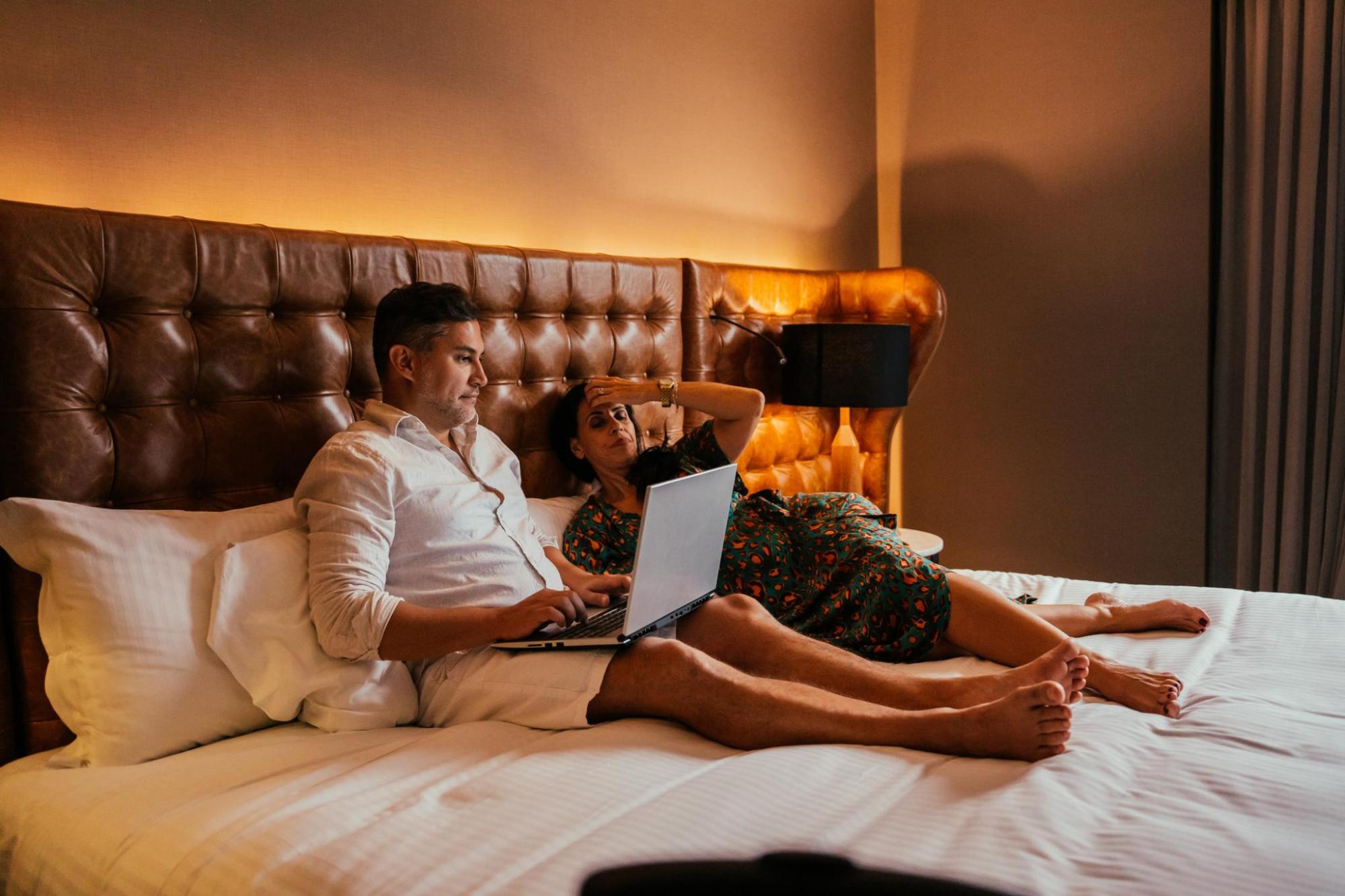 Couple on a hotel bed, looking at a laptop in a warmly lit room