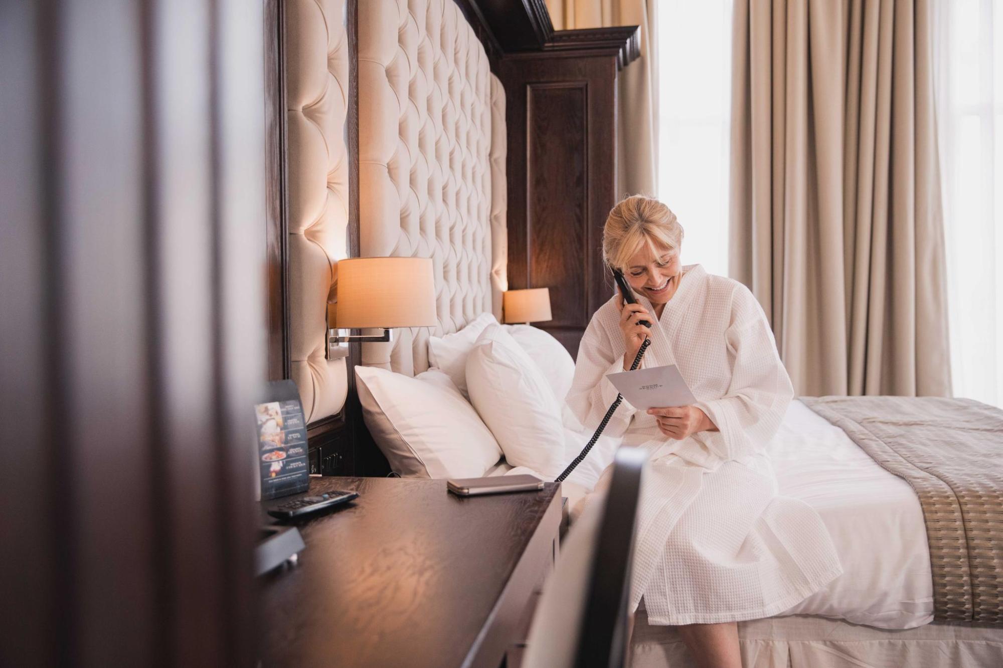 Woman in a hotel robe sits on a bed, smiling while speaking on the room phone and reading a menu