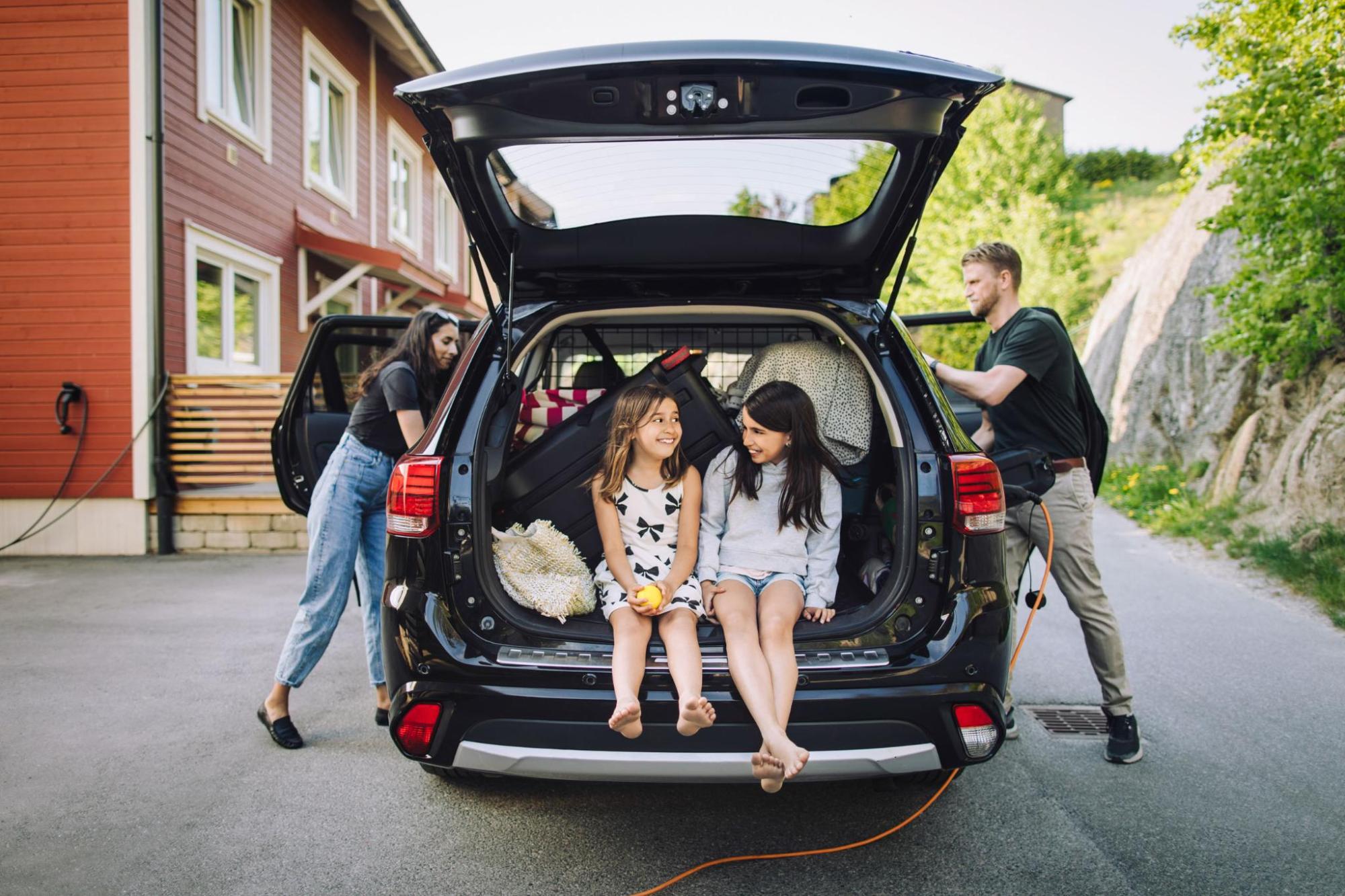 Two adults pack luggage into a car while two children sit in the boot