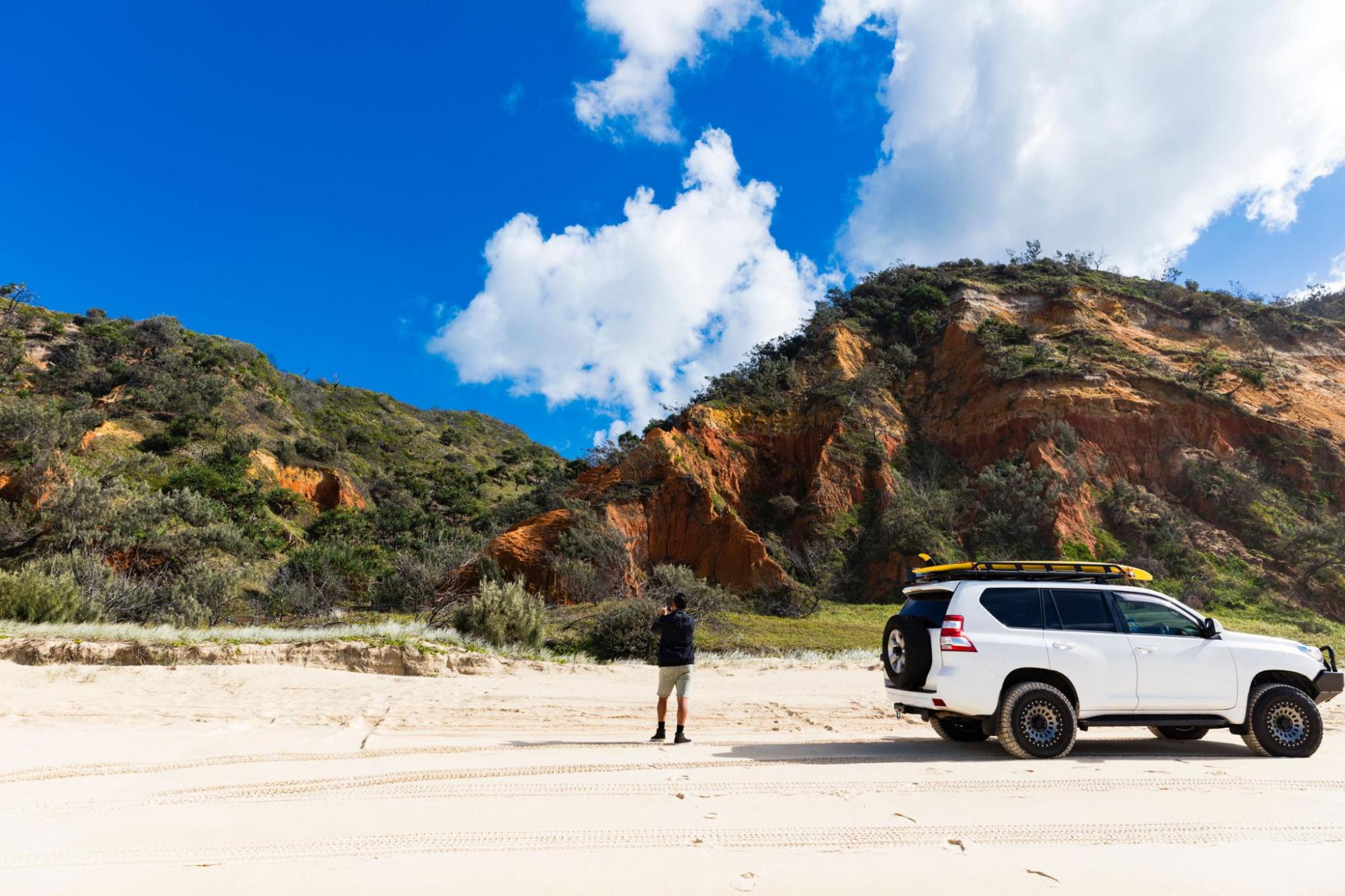 A person stands next to a white SUV on a beach with rolling hills in the background