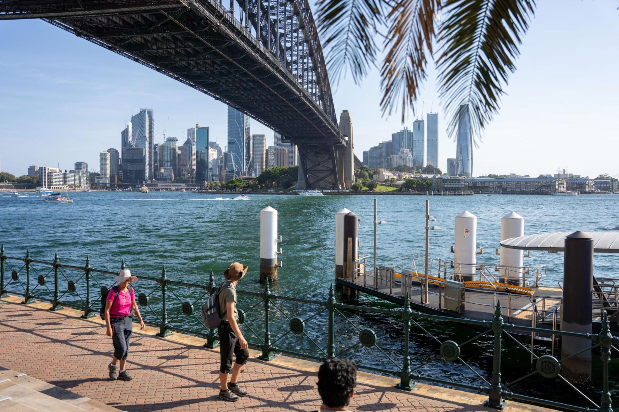 Two people walk along a waterfront path while a ship passes under a bridge, with skyscrapers in the background.