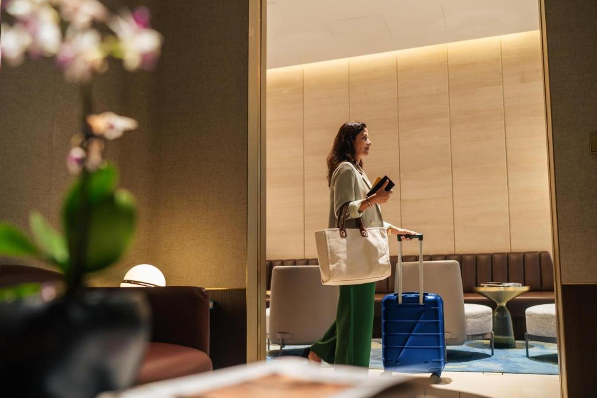 Frau mit weißer Tasche und blauem Rollkoffer steht in modern eingerichteter Hotellobb