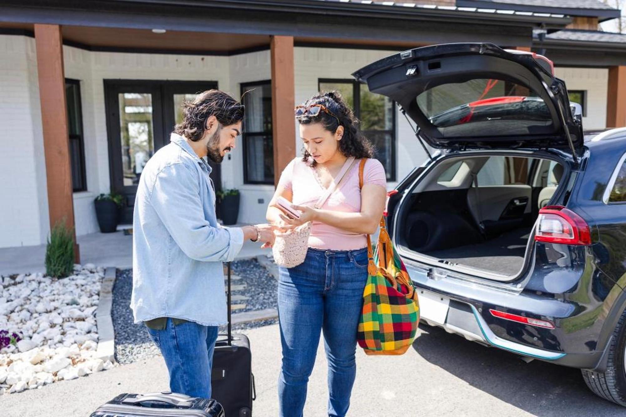 Two travellers stand beside an open car boot outside a house, checking a phone with luggage nearby