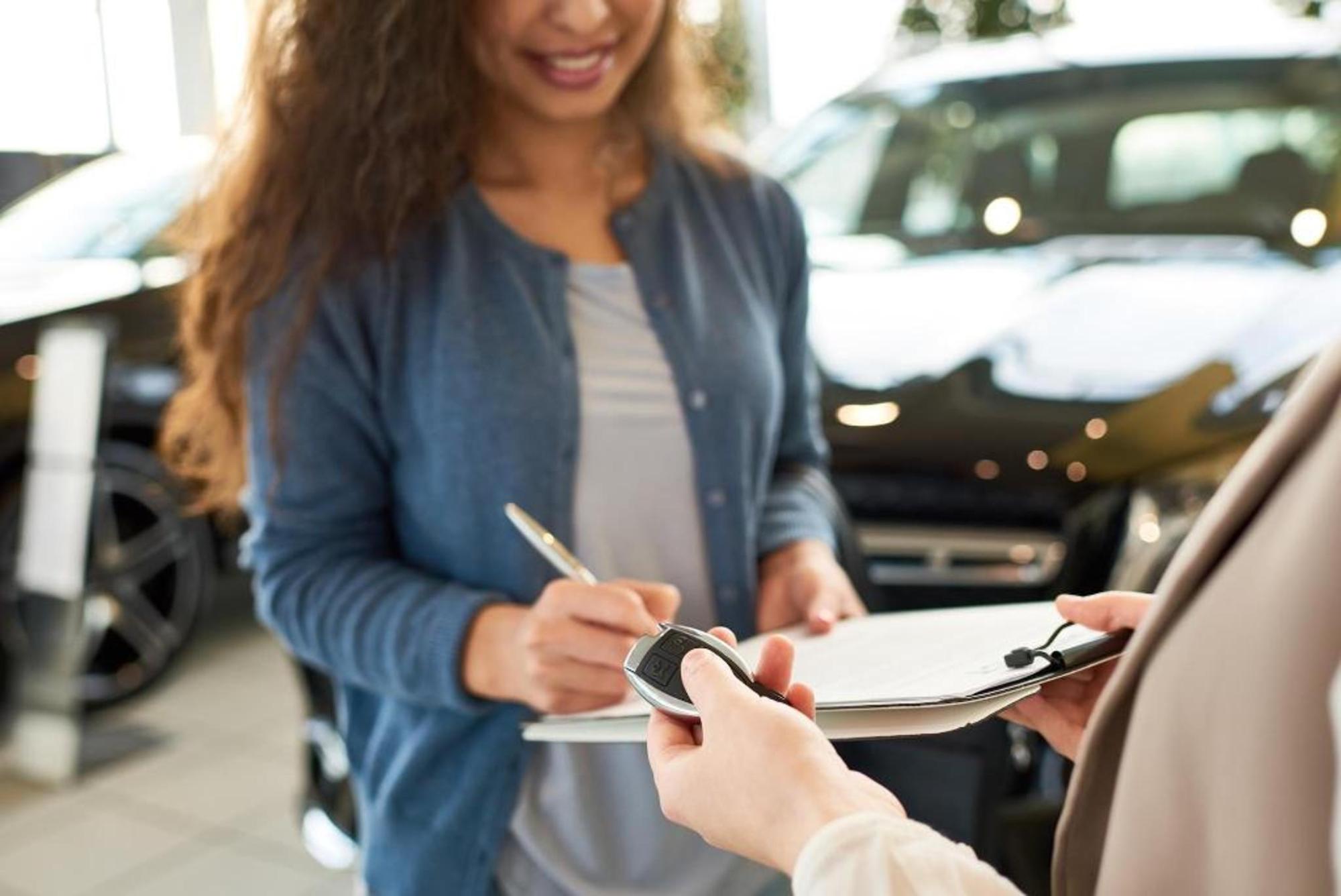 Two people exchange car keys and paperwork beside a car in a showroom