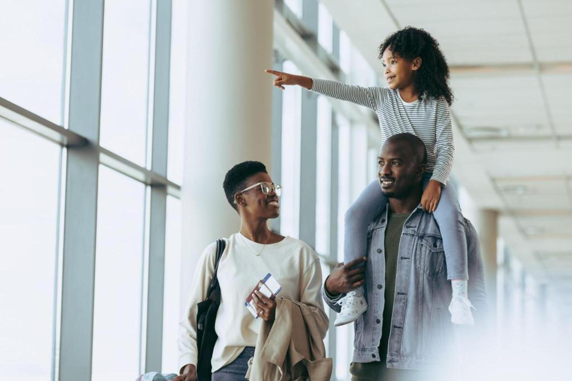 A family walks through an airport terminal, with a child sitting on a man’s shoulders and pointing ahead