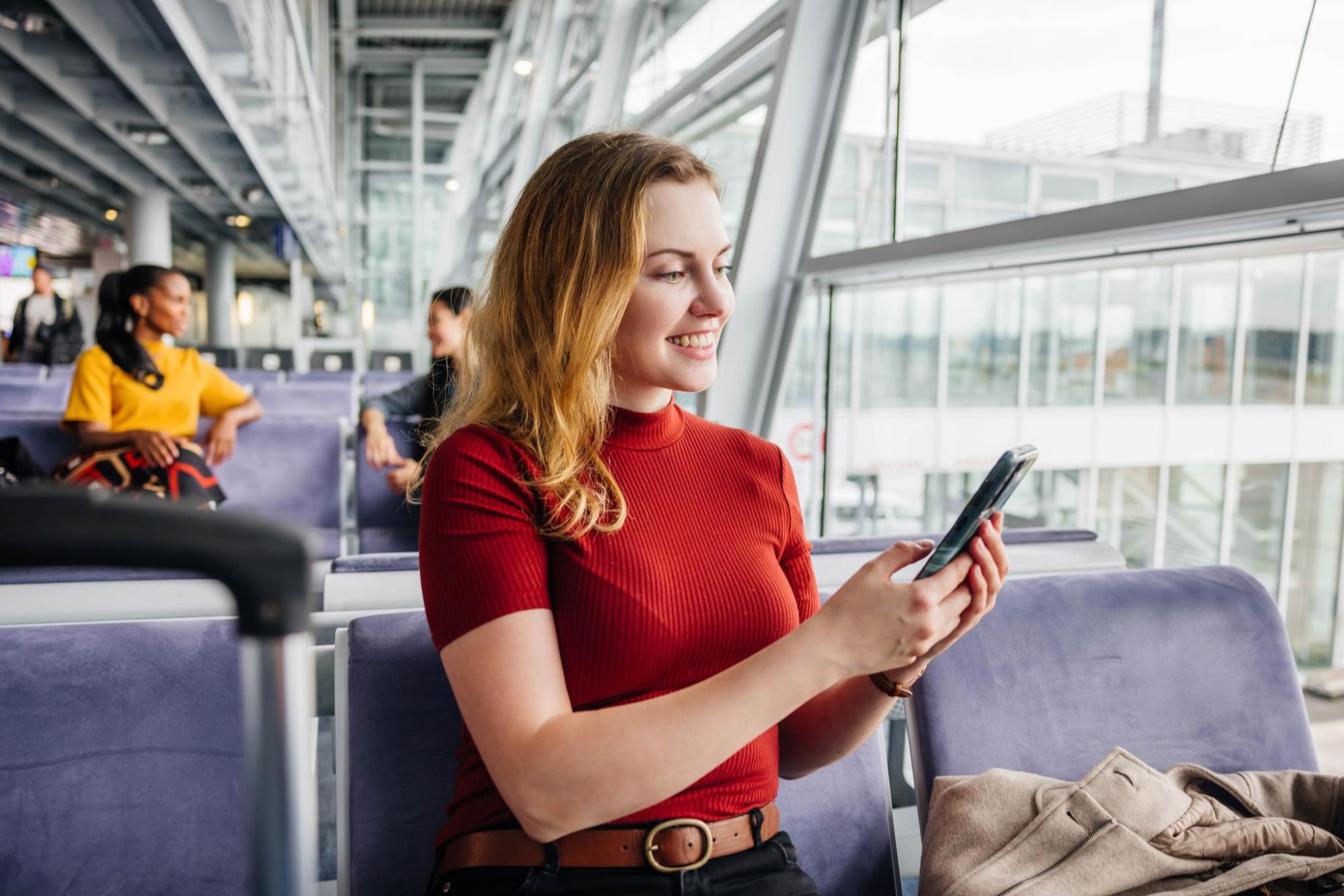 Une femme souriante sur son téléphone est assise sur une chaise d'aéroport