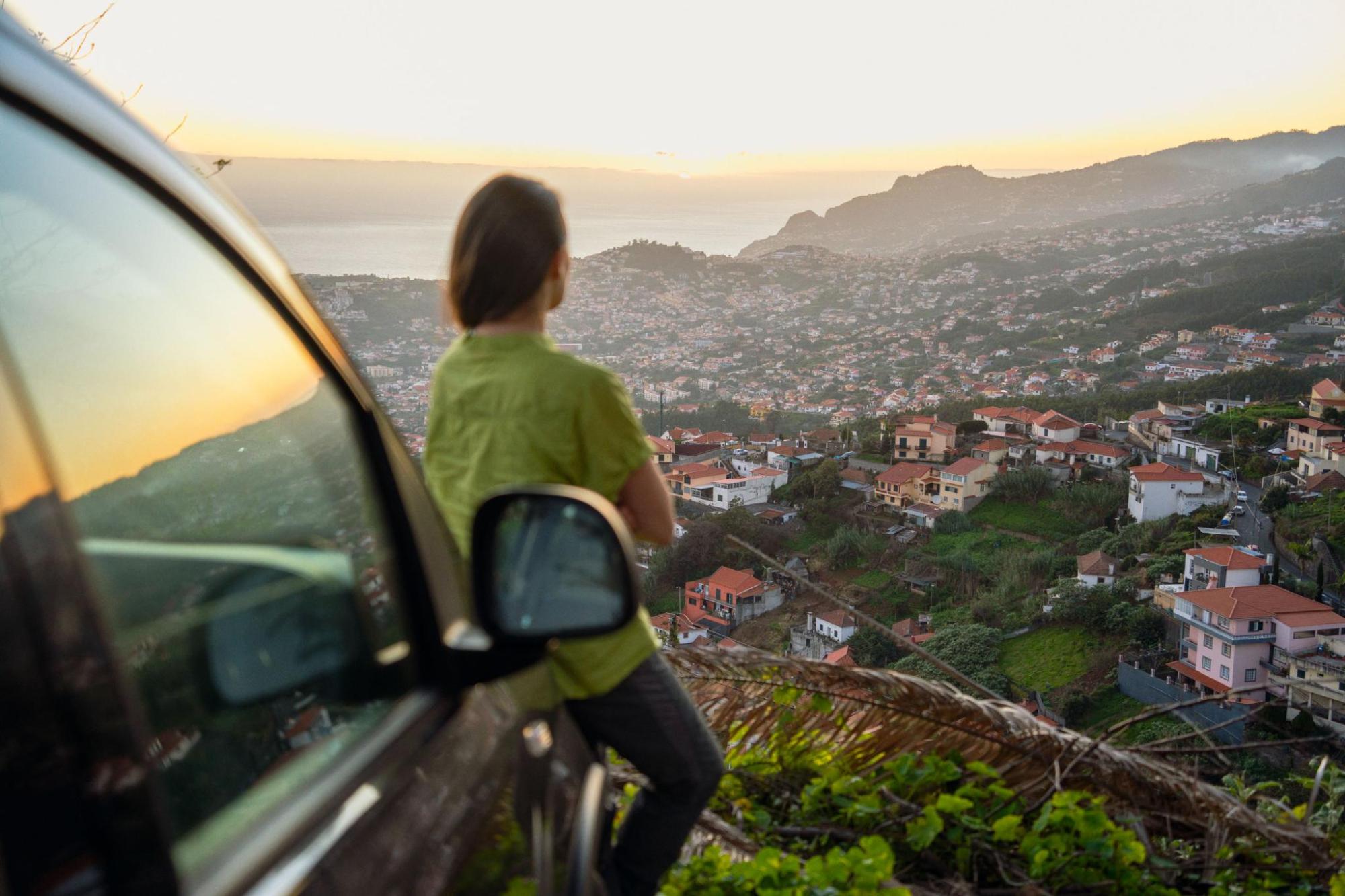 A person sits on the bonnet of a car and looks over a coastal hillside town 