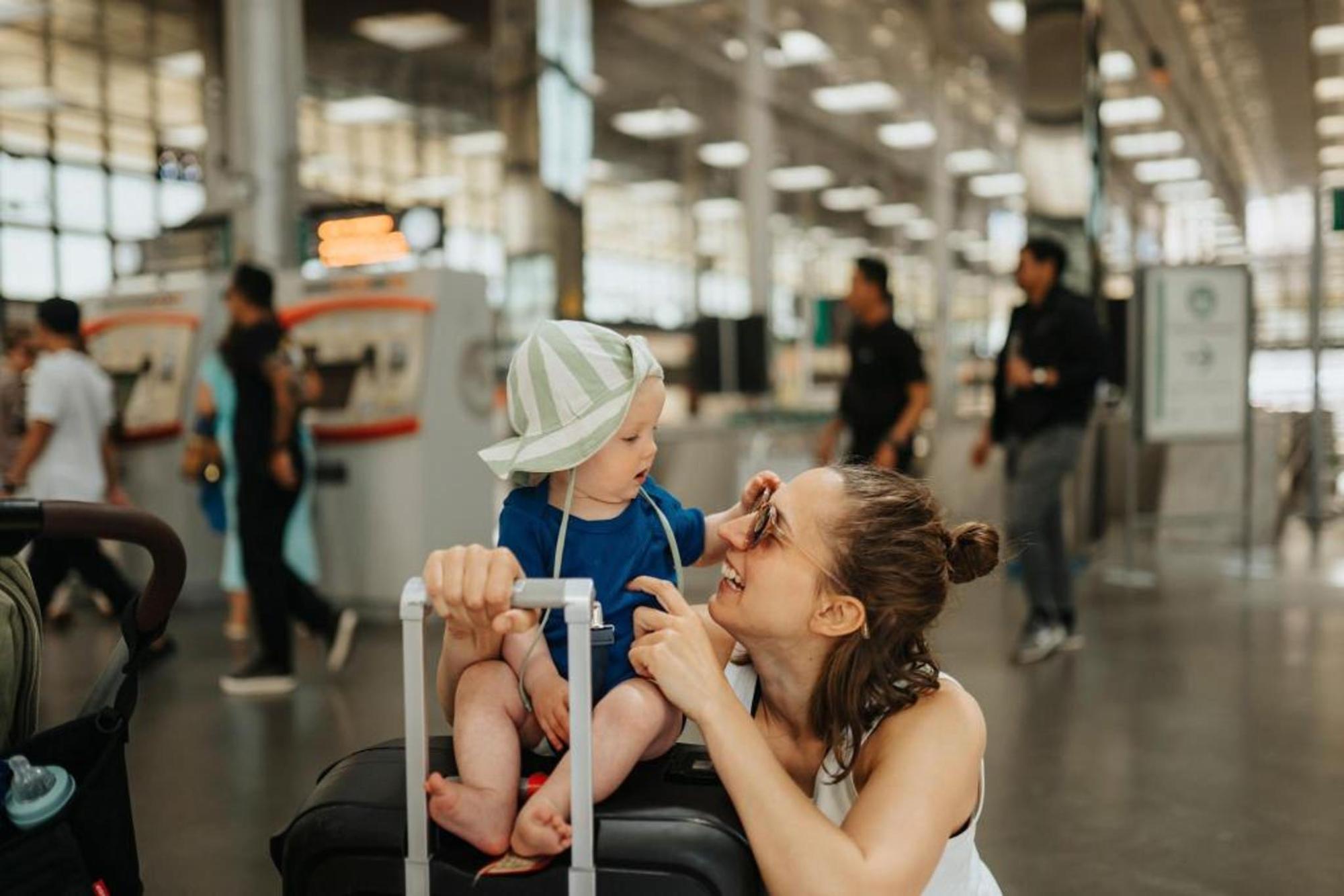 A woman smiling at a toddler sitting on a suitcase in an airport terminal