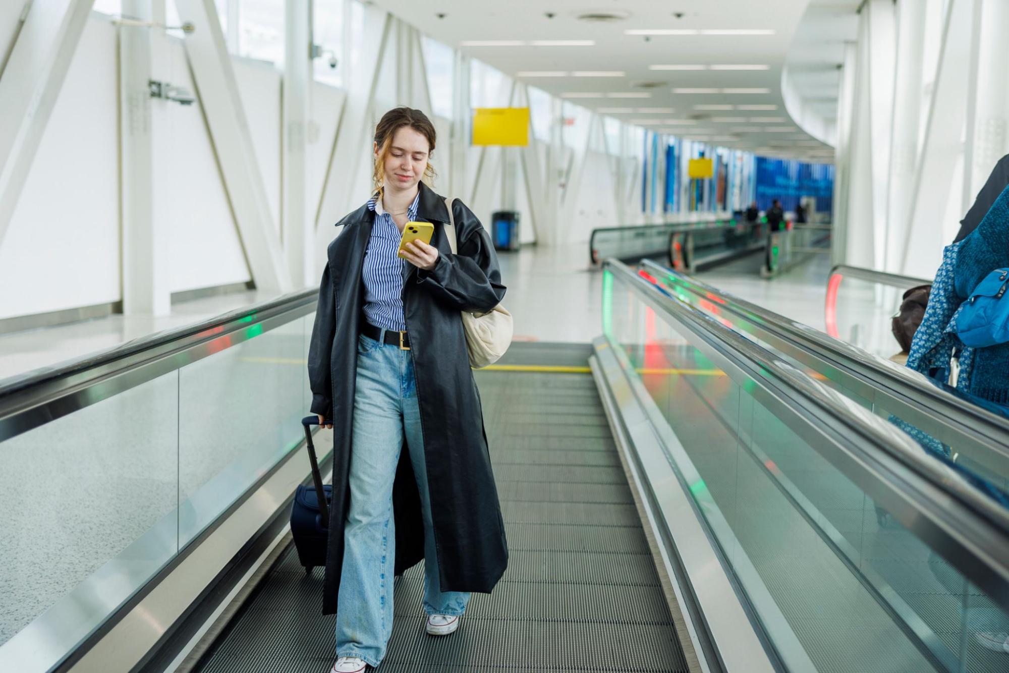Frau mit langem schwarzem Mantel und blauer Jeans geht auf einem Flughafentransportband und schaut auf ihr Handy