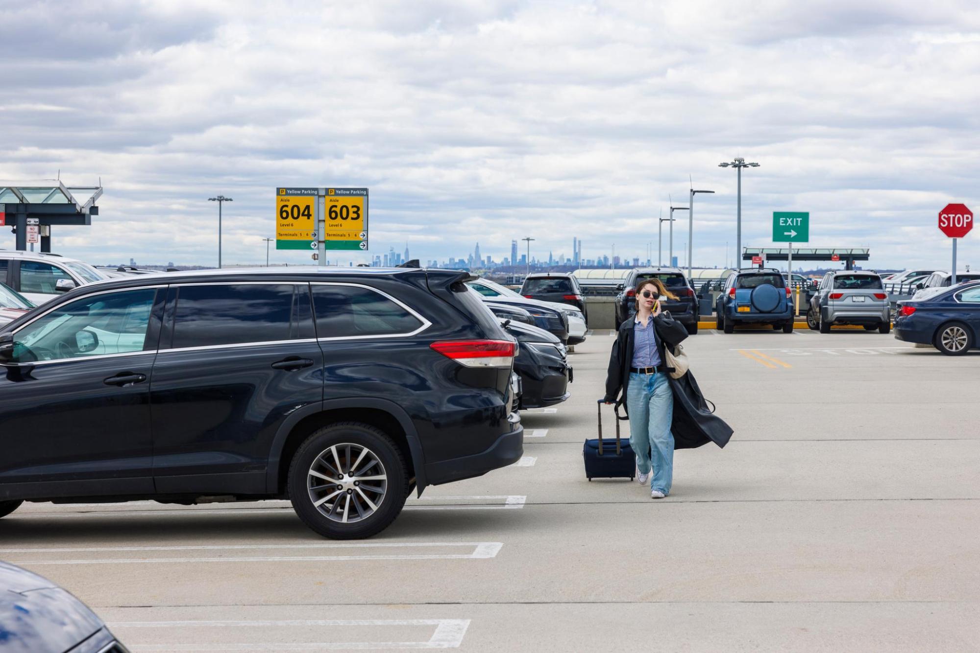 Une jeune femme sur le parking d’un aéroport, transportant une valise, un téléphone à l’oreille, entourée de voitures et de panneaux de signalisation