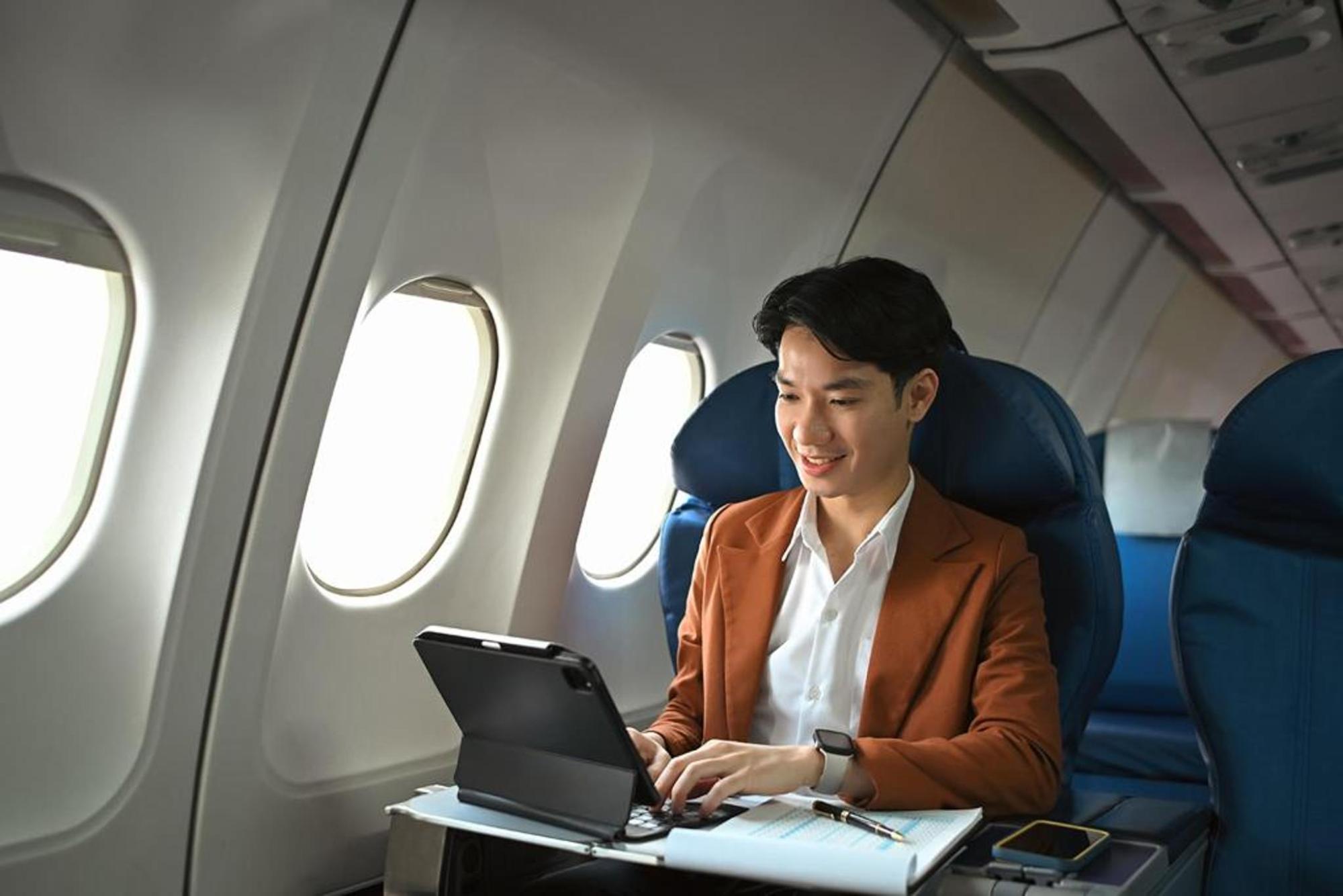 Ein Mann sitzt an einem Fensterplatz im Flugzeug und tippt lächelnd auf einem Tablet.