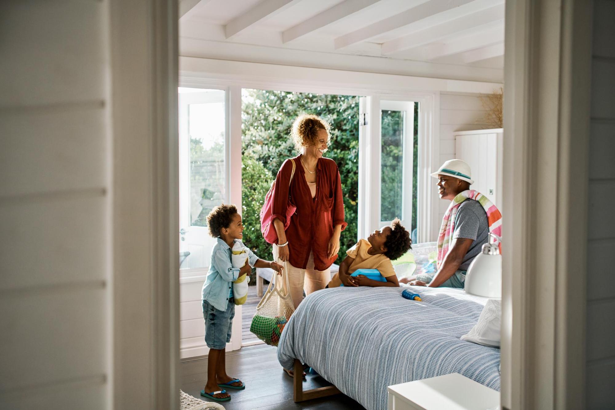 Familie in einem hellen Schlafzimmer, zwei Kinder, eine Frau mit rotem Oberteil und ein Mann mit Hut sitzen und stehen um ein Bett