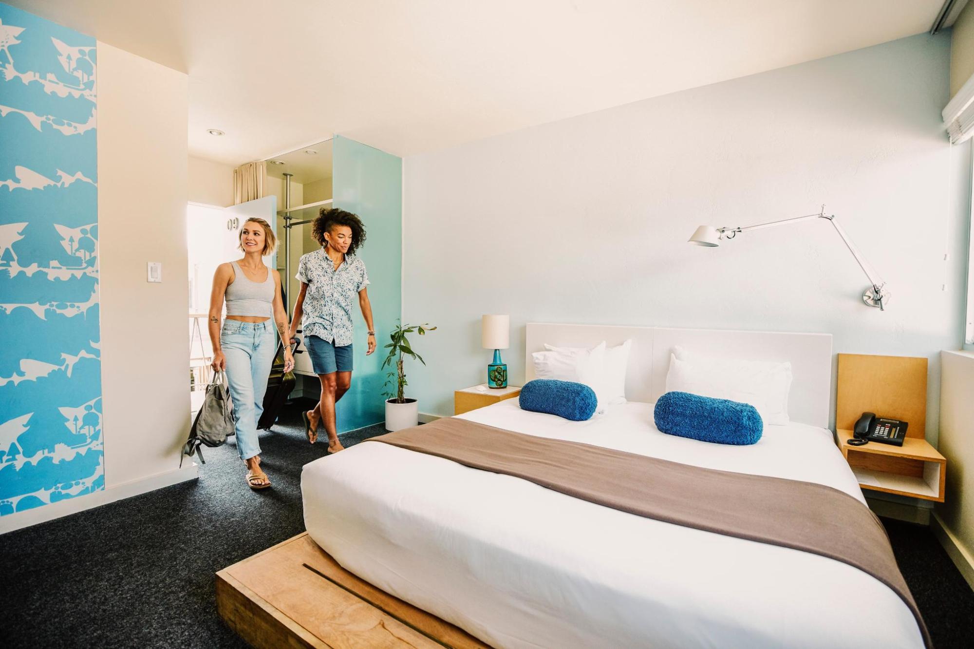 Zwei Frauen betreten ein helles Hotelzimmer mit großem Bett, blauen Kissen und moderner Einrichtung.
